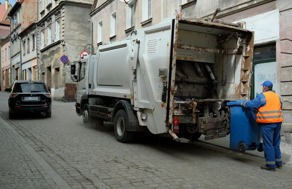 A sanitation worker wearing a blue uniform and an orange safety vest is standing on a cobblestone street next to a large white waste collection truck, positioned with its rear hopper open and facing the curb. The worker is holding a blue wheeled rubbish bin, preparing to empty it into the truck's compactor, which is partially visible with rusted metal parts inside. The scene is set in an urban area, with multi-storey residential buildings featuring brick and stone facades lining the street. Parked cars, including a black hatchback, are visible along the roadside. The street appears to be a designated waste collection route, with signage indicating no parking zones. This image, provided by Man with Van Freezywater, illustrates the process of home waste removal within a property clearance or moving-related context, highlighting the practical steps involved in rubbish disposal during a house removal or relocation service.