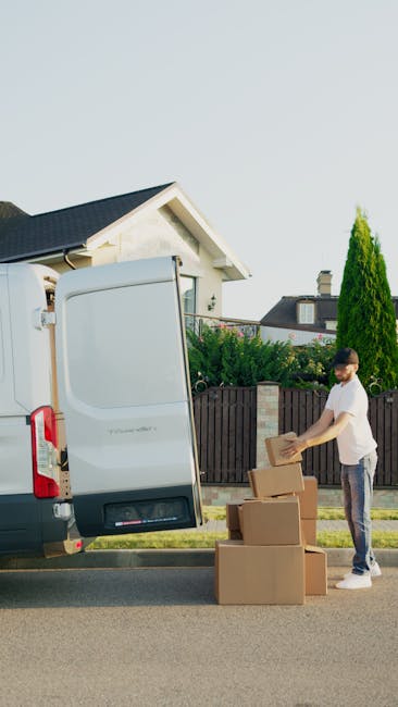 A young man wearing a white t-shirt and jeans is standing on the pavement beside a white panel van with its rear doors open. He is handling a cardboard box, part of a stack of similar boxes, which are
