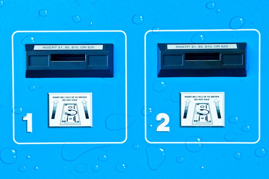 Close-up of a bright blue coin-operated laundry payment panel with two separate slots labeled '1' and '2' for inserting coins or bills, with instructions for payment options displayed above each slot. The panel features a waterproof surface with visible water droplets, indicating it is situated outdoors or in a humid environment. The payment panel is part of a building or laundry service area, relevant to home relocation or packing processes. Natural lighting highlights the clean, industrial design of the coin slots and instructions, which are clear and legible. This image represents the typical payment equipment used in laundry facilities, often involved in house moves or logistics when providing laundry or cleaning services as part of a moving process.
