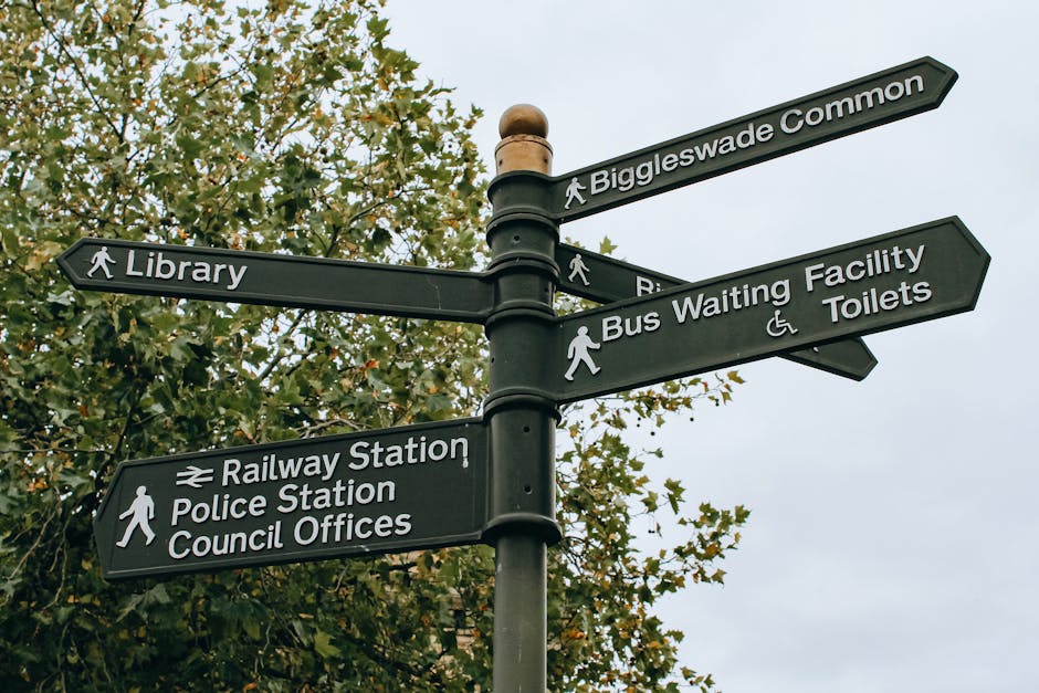 A black multi-directional pedestrian signpost situated outdoors against a backdrop of green leafy trees and a cloudy sky. The sign features several labels including 'Biggleswade Common' pointing to the upper right, 'Library' pointing left, 'Bus Waiting Facility and Toilets' pointing right, and 'Railway Station, Police Station, Council Offices' pointing downward. The signs are attached to a central pole, which is topped with a decorative spherical finial. The scene likely depicts an area near community amenities, suitable for navigating through different parts of the town or area, relevant to relocation or travel logistics, with [COMPANY_NAME] providing related removals and transportation services.