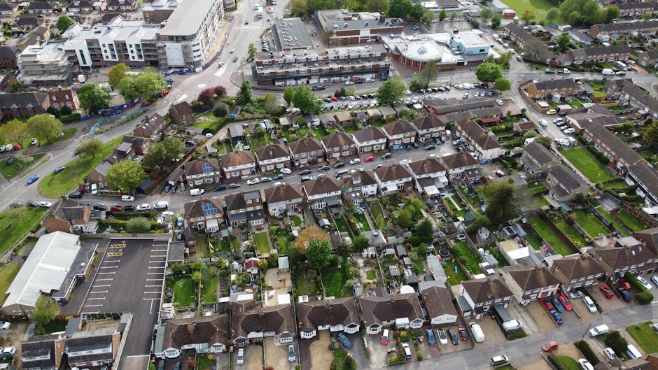 An aerial view of a residential area showing rows of terraced houses with small, well-maintained gardens and driveways, some equipped with solar panels on the roofs. The street is lined with parked cars, and a few vehicles are visible on the road. Adjacent to the houses, there is a large parking lot for a commercial or community building, with marked parking spaces. In the background, some larger modern buildings and green spaces can be seen, along with trees scattered throughout the area. The scene captures a typical suburban neighbourhood within Enfield, suitable for house removals and home relocation services by Man with Van Freezywater, illustrating the typical environment encountered during furniture transport and packing and moving processes.