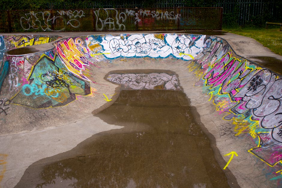 A concrete skate park bowl with curved walls covered in colourful graffiti tags and murals, situated outdoors under diffused daylight. The surface of the bowl features patches of dark wet concrete, indicating recent watering or cleaning, with visible footprints and scuff marks. Surrounding the bowl is a flat concrete area with some scattered paint splashes, leading to a paved pathway that encircles the park. In the background, there is a rusted metal fence adorned with additional graffiti, and beyond that, a grassy area with trees and shrubbery. The scene suggests an urban recreational space used for skateboarding or parkour activities, with no people present. The image provides a detailed view of the textured, graffiti-adorned surface and the outdoor environment typically associated with community skate parks, pertinent to services involving equipment transport or setting up for outdoor activities by companies like Man with Van Freezywater.