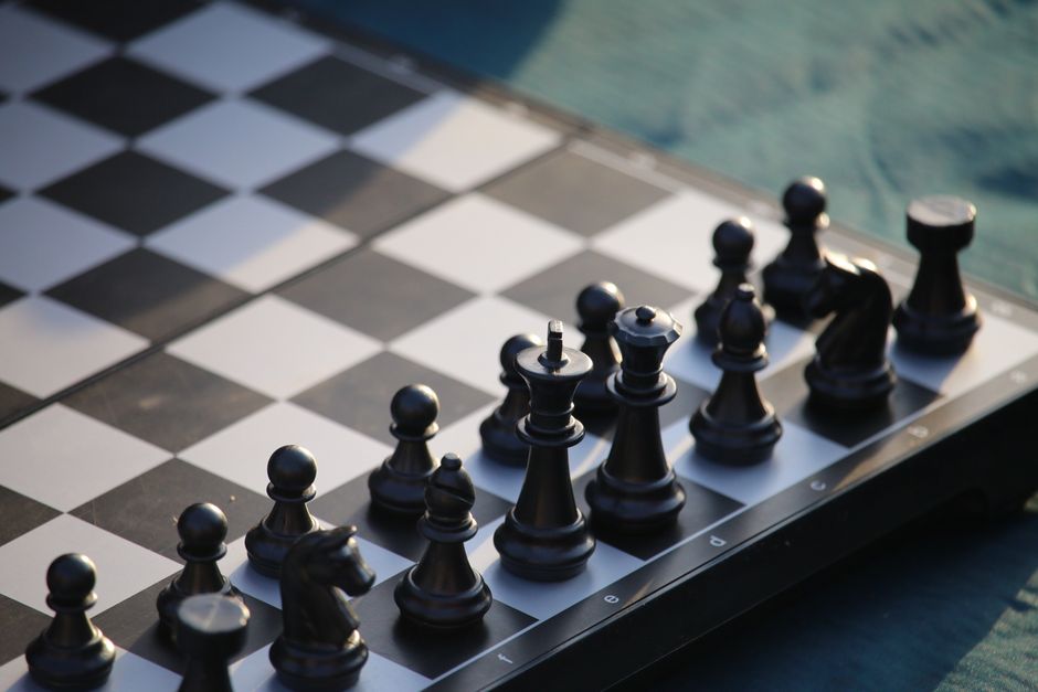Close-up view of a standard chessboard positioned on a surface near a window, with black chess pieces arranged in their starting positions. The pieces include pawns, rooks, knights, bishops, queens, and kings, made of plastic or wood, with the black pieces on light and dark squares. The background shows a blurred view of water outside, suggesting the scene might be set near a window overlooking a body of water. Inside, some packing materials like paper or thin padding may be visible around the chessboard area. This scene captures an organized arrangement of the chess set in a well-lit environment, with natural daylight illuminating the objects, consistent with a setting where packing or unpacking could be happening during a home relocation process, as seen in professional removals services.