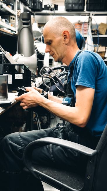 A man with a shaved head and light skin, wearing a blue t-shirt, is seated on a black office chair inside a workshop or storage area filled with shelves holding various tools, electronic devices, and packaging materials. He is focused on holding and examining an electronic component or device with both hands, surrounded by cables, small boxes, and equipment. The environment appears to be well-lit and organized for technical or repair work, which is relevant to home relocation services involving packing, disassembly, or preparation of items for moving, as provided by Man with Van Freezywater.