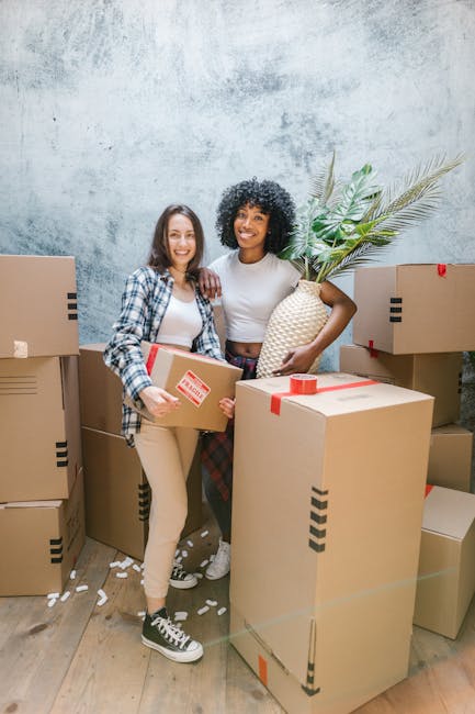 Two women stand inside a room surrounded by stacked cardboard boxes during a home relocation process, with one holding a box and the other holding a large woven basket with green tropical leaves. The boxes are sealed with red tape and are of various sizes, positioned on the wooden floor and behind them. The women appear to be preparing for or engaged in packing and moving activities, with one wearing casual attire and the other in a plaid shirt over a white top. The room has a textured, light grey wall and is well-lit, reflecting an organized packing process. This image exemplifies the logistical aspects of furniture transport and packing as part of a local removals service, similar to those offered by Man with Van Freezywater, with the focus on loading and handling household items.