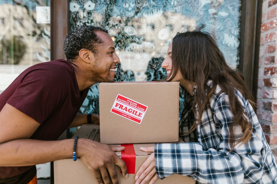 A person wearing a dark blue t-shirt with a patterned pocket is placing a clear glass drinkware item into a cardboard box filled with white packing peanuts for protection. The box, labelled with a red sign indicating 'Handle with Care - Glass,' is situated indoors on a wooden floor, near a window that allows natural light to illuminate the scene. In the background, there are additional cardboard boxes, some sealed and stacked, indicating the process of packing for a home relocation or moving project. The person’s arms are visible, and they are carefully handling the fragile item to ensure safe transport, which is typical of furniture transport and packing during house removals. The image conveys the careful packing of glassware as part of the logistics involved in professional removals, supported by the presence of packing materials, protective packaging, and a focus on safe handling during the moving process.