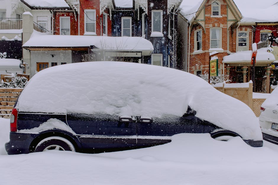 A black hatchback car parked on a snow-covered street in Freezywater, with its roof and windows heavily blanketed in snow, partially obscuring the vehicle. The car is situated near a pavement and adjacent to other parked vehicles. In the background, there are residential houses with varying architectural styles, some with brick facades and bay windows, also covered in snow. The snow is fresh and abundant, creating a thick layer on the car and surrounding area, with snowflakes visibly falling. The scene depicts a winter setting during a snowstorm, and the car appears to be stationary, possibly awaiting a home relocation or moving process by a professional removals service like Man with Van Freezywater, who manage loading and transport in winter conditions.