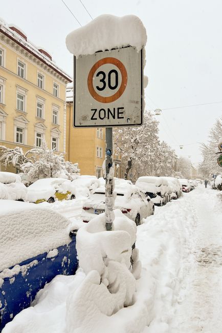 A snow-covered street scene featuring a traffic sign indicating a 30-zone, with the sign and its supporting post heavily blanketed in snow and ice. Parked cars along the street are also covered in snow, with their windows, roofs, and side panels coated. In the background, multi-storey buildings with yellow and beige facades are partially visible through the wintry atmosphere, and snow-laden trees line the sidewalk. The snow on the ground and parked vehicles suggests recent snowfall. This setting may depict an urban environment where proper parking and clearance are important for home relocations or moving services. The snow accumulation around the sign and cars highlights the necessity for careful planning during winter moves, as managed by companies like Man with Van Freezywater. The overall scene demonstrates the typical conditions faced when carrying out furniture transport or home relocation activities in snowy weather conditions, emphasizing the importance of weather-aware logistics for removals companies.