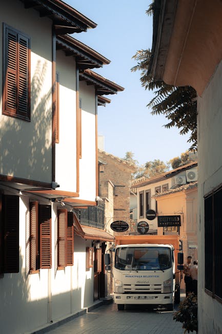 A small white moving van with a flat front is parked on a narrow street between two buildings in a residential area during daytime. The van is positioned facing towards the viewer, with its driver's side visible, and appears to be in the process of loading or unloading household furniture and boxes. To the left, there is a two-storey building with white walls, brown window shutters, and a tiled roof, while on the right, another building with similar architectural features is partially visible. In the background, additional structures and a clear blue sky are visible, indicating bright weather. The scene reflects a home relocation or furniture transport process, with the company Man with Van Freezywater potentially involved in the moving services. This environment represents a typical urban setting for house removals, with a focus on loading and transportation logistics.