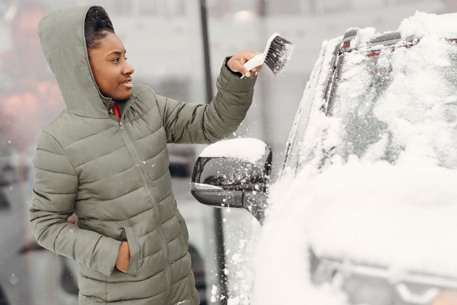 A woman wearing a light grey winter jacket with a hood is seen standing outside in front of a snow-covered vehicle, likely a van used for house removals. She is holding a brush and cleansing the snow from the side mirror of the van, which is partially covered in snow on the windshield and body. The background shows a blurred outdoor setting, suggesting cold weather conditions typical of winter. Inside the property, visible through an open doorway, there are cardboard boxes and various packing materials, including plastic wrap and bubble wrap, indicating preparations for a home relocation or moving process. The vehicle is parked on a paved area, ready for loading or transportation, and the setting reflects the packing and moving activities managed by [COMPANY_NAME], focusing on professional furniture transport and home removals in freezing weather.