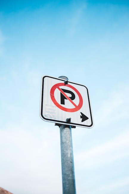 A rectangular white parking restriction sign mounted on a metal pole against a blue sky with some clouds. The sign displays a black letter 'P' crossed out with a red circle and slash, indicating no parking, and includes a black arrow pointing to the right at the bottom. In the context of house removals and relocation services, such signage is relevant for locating designated parking areas for delivery and moving vehicles. Man with Van Freezywater, a professional removals company, would consider parking restrictions like this when planning home relocation and furniture transport. The sign's placement and visibility are crucial for ensuring smooth loading processes, such as loading furniture, boxes, or appliances into a moving van. The image highlights the importance of understanding parking regulations to facilitate efficient packing and transport during house moves, particularly in urban or restricted parking zones.