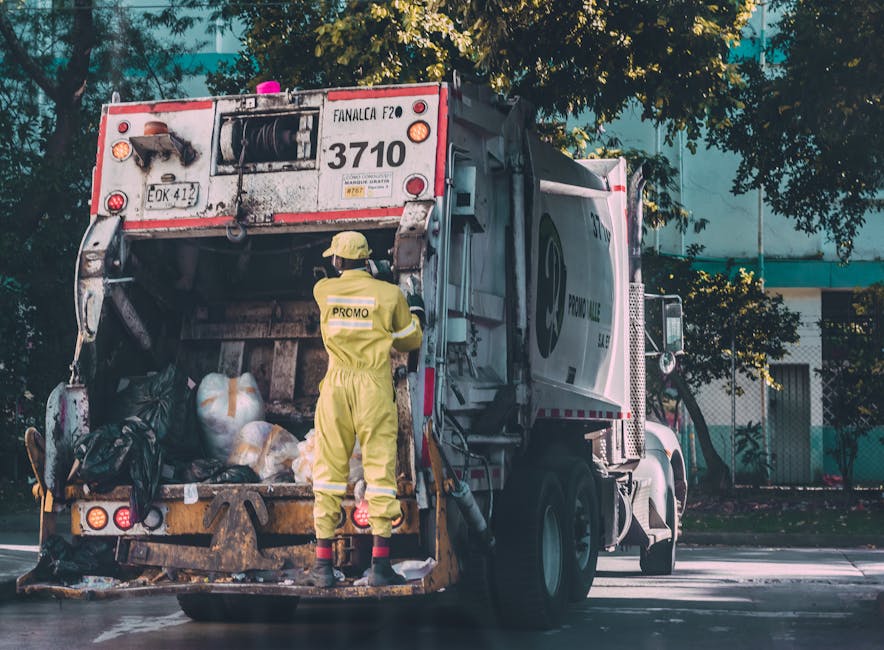A waste collection vehicle with the rear hatch open, showing black and white waste bags and loose rubbish inside, is parked on a street near a sidewalk. A sanitation worker dressed in a yellow protective suit, including a helmet and gloves, stands at the back of the truck, using a tool to manage the waste. The truck is branded with the logo of a waste management service and has visible identification numbers and markings. The environment is outdoors, with trees and a building with windows in the background, and sunlight illuminating the scene. This image relates to waste disposal processes handled by professional services like Man with Van Freezywater, a company specialising in house removals and property transfers, including the careful transport of packing materials and cleaning during home relocation or moving logistics.
