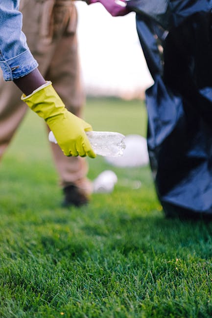 A person wearing yellow rubber gloves is placing a clear plastic bottle into a recycling bin during a home relocation cleanup outside a property. The scene captures a section of the front garden with well-maintained green grass and a partially visible black recycling container. In the background, another individual, dressed in neutral colours, is seen wearing beige trousers and brown shoes, standing on the pavement near the entrance. The image emphasizes the importance of proper waste disposal and recycling as part of the house removal process, consistent with the services offered by Man with Van Freezywater for moving and packing logistics.