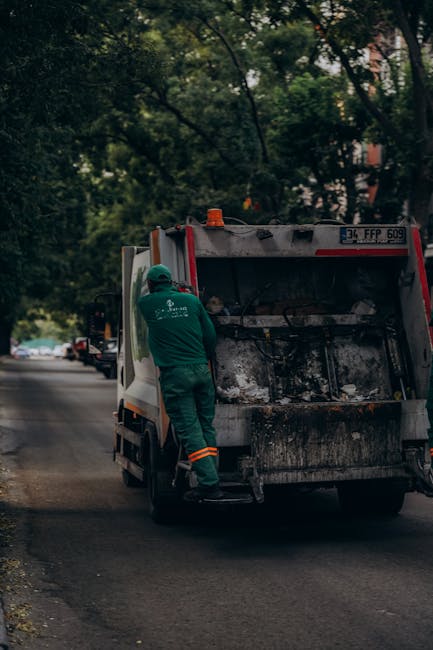 A waste collection vehicle with the rear hatch open, showing black and white waste bags and loose rubbish inside, is parked on a street near a sidewalk. A sanitation worker dressed in a yellow protective suit, including a helmet and gloves, stands at the back of the truck, using a tool to manage the waste. The truck is branded with the logo of a waste management service and has visible identification numbers and markings. The environment is outdoors, with trees and a building with windows in the background, and sunlight illuminating the scene. This image relates to waste disposal processes handled by professional services like Man with Van Freezywater, a company specialising in house removals and property transfers, including the careful transport of packing materials and cleaning during home relocation or moving logistics.